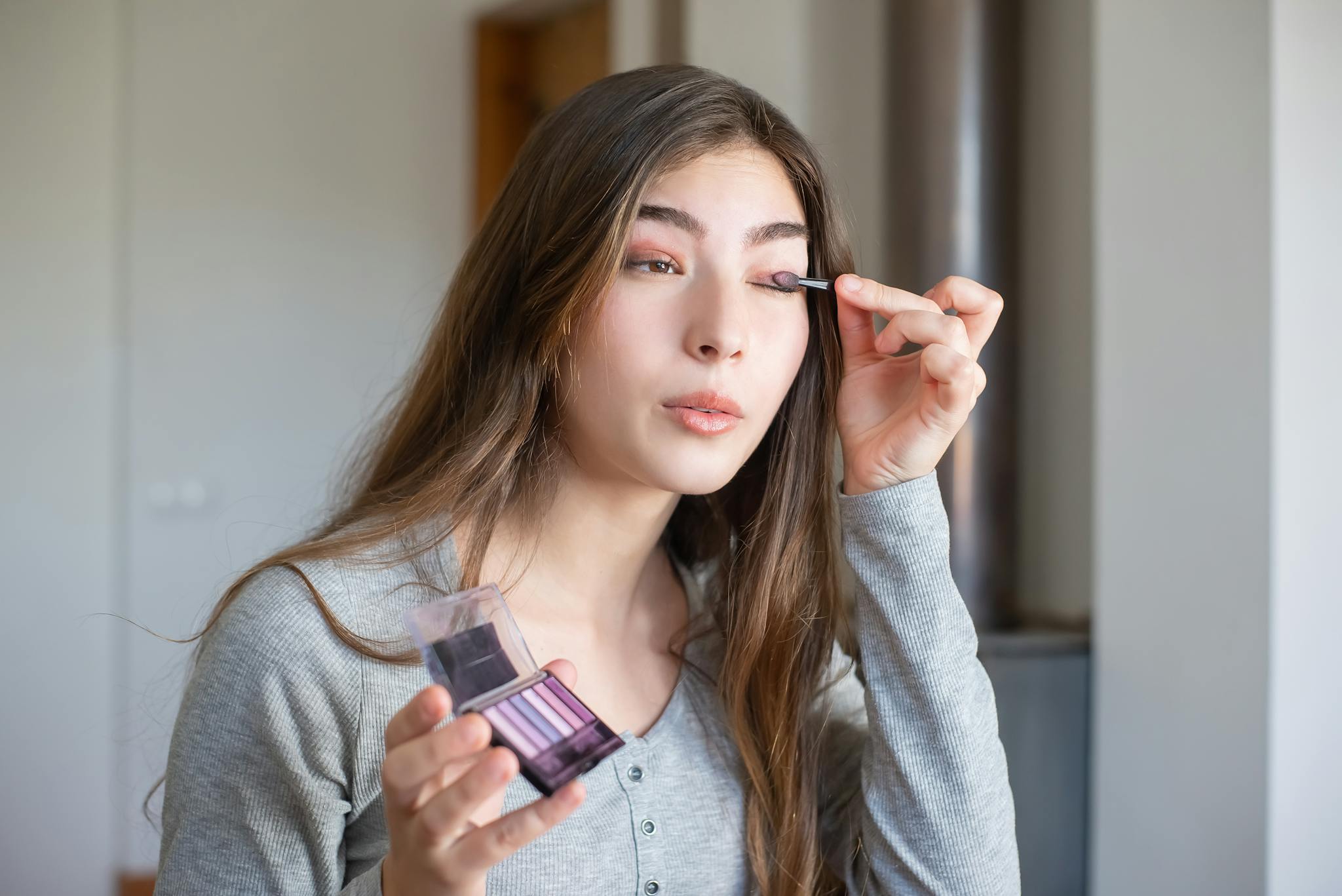 Asian woman applying makeup with eyeshadow palette indoors, showcasing beauty routine. in home page section.